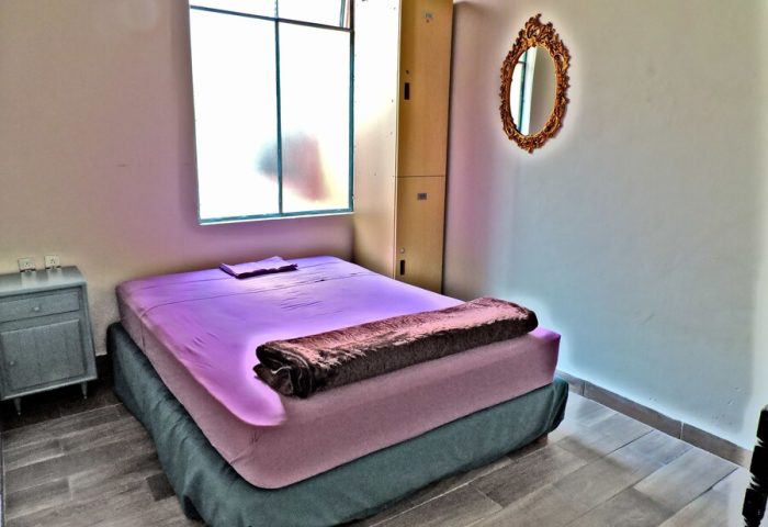 Capsule Hostel room: bed with purple sheets, brown blanket, lockers, and mirror. Gay-friendly hotel for gay men travel advice.