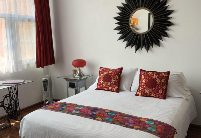 Casa Comtesse gay-friendly hotel room. Bed with colorful runner, decorative pillows, and sunburst mirror. Red accents. Cozy, stylish.