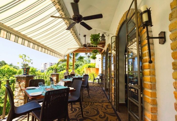 Casa Cupula, gay-friendly hotel. Outdoor dining area with tables set for a meal. Tropical plants and trees in the background. Ceiling fans above.