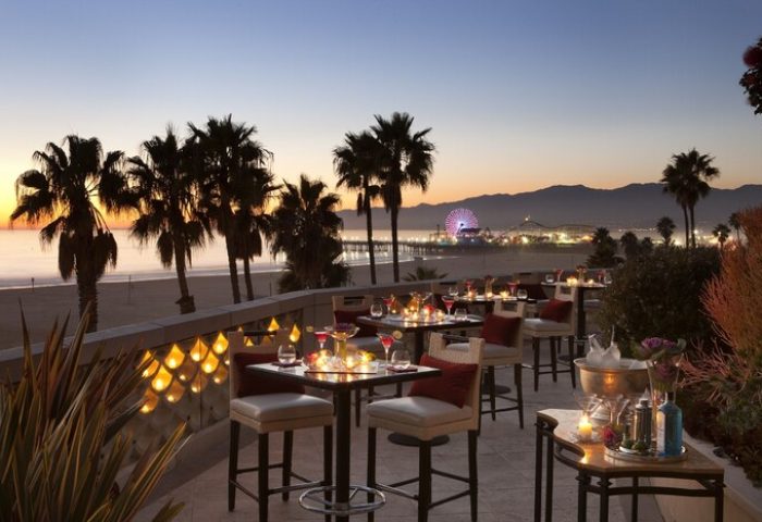 Casa Del Mar gay-friendly hotel outdoor dining area at sunset. Palm trees, beach, and Santa Monica Pier views. Upscale travel for gay men.