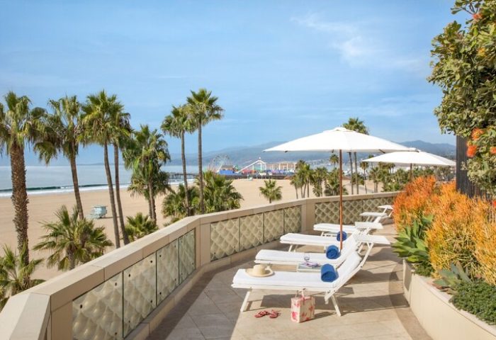 Casa Del Mar gay-friendly hotel view. Beachfront with palm trees, lounge chairs, umbrellas. Santa Monica Pier visible. Perfect gay travel spot.