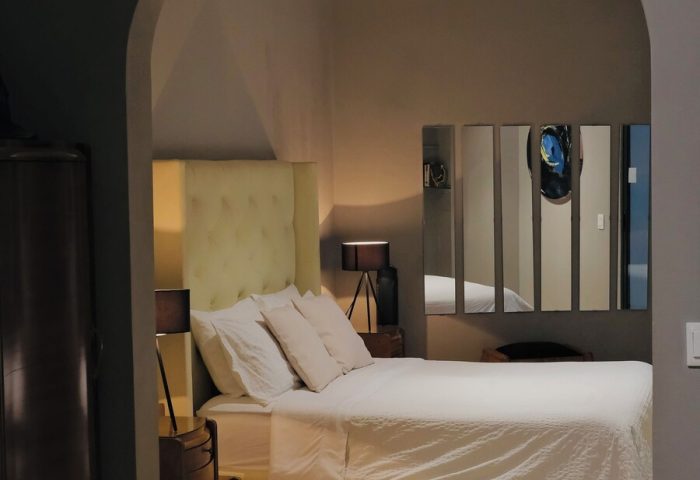 Casa Mannach gay-friendly hotel bedroom. Archway view of bed with white linens, tufted headboard, and mirrored wall art. Interior design.