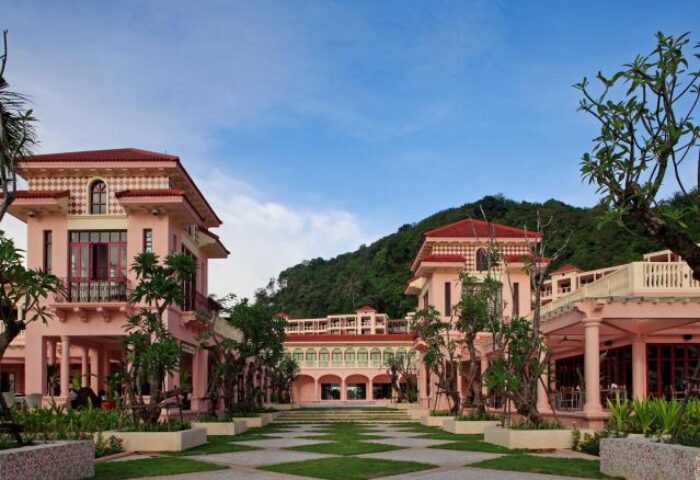 Centara Grand Beach Resort Phuket view. Gay-friendly hotel with pink buildings, green lawn, and mountain backdrop. A serene gay travel destination.