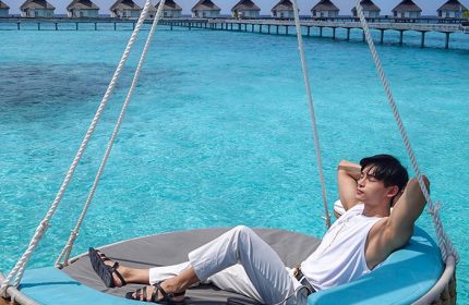 Gay travel: man relaxes on a swing at Centara Grand Island Resort & Spa, Maldives. Overwater bungalows in the background.
