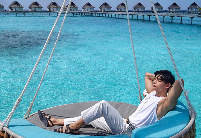 Gay travel: man relaxes on a swing at Centara Grand Island Resort & Spa, Maldives. Overwater bungalows in the background.