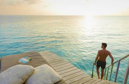 Gay-friendly Centara Ras Fushi Resort & Spa Maldives: Man entering turquoise water from a private deck at sunset. Ideal gay travel destination.