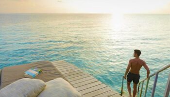 Gay-friendly Centara Ras Fushi Resort & Spa Maldives: Man entering turquoise water from a private deck at sunset. Ideal gay travel destination.