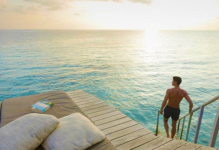 Gay-friendly Centara Ras Fushi Resort & Spa Maldives: Man entering turquoise water from a private deck at sunset. Ideal gay travel destination.