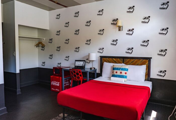 Chicago Getaway Hostel room. Gay-friendly hotel with bed, desk, and Polaroid camera decals. Chicago flag pillow. Red Coca-Cola mini fridge.
