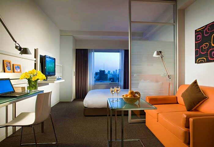 Citadines Shinjuku Hotel room. Gay-friendly hotel with city view, desk, laptop, orange couch, and champagne. Perfect for gay men's travel in Shinjuku.