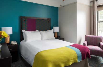 City Suites Chicago gay-friendly hotel room. Queen bed with colorful blanket, headboard, & accent wall. Chair by window. LGBTQ travel.
