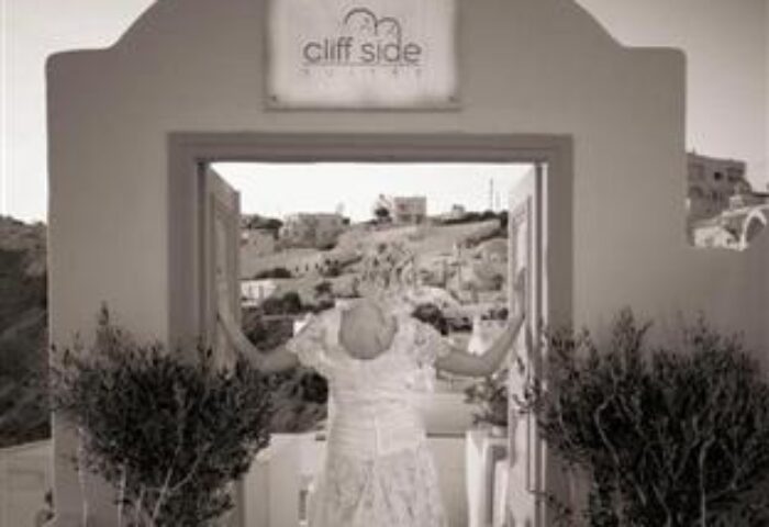 Cliff Side Suites gay-friendly hotel view. Woman in white dress opens doors to a balcony overlooking a village. Sepia tone.