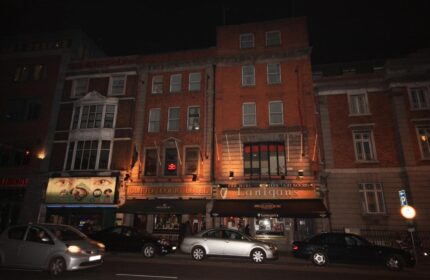 Night view of Clifton Court Hotel, a gay-friendly hotel in Dublin. The hotel's facade is visible, with cars parked in front.