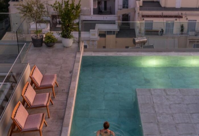 Rooftop pool at Coco-mat Athens BC, a gay-friendly hotel. Woman swims, chairs nearby. Athens cityscape in background. Ideal for gay travel.