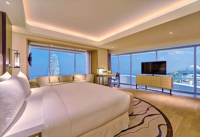 Conrad Manila gay-friendly hotel room. Spacious suite with a large bed, desk, and floor-to-ceiling windows offering ocean views and a Ferris wheel.