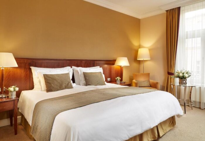 Corinthia Hotel Budapest bedroom. Gay-friendly, pet-friendly hotel with a king-size bed, bedside tables, lamps, flowers, chair, and window.