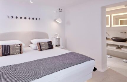 Cosmopolitan Suites gay-friendly hotel room. Minimalist design with white walls, bed with decorative pillows, and bathroom view. Elegant travel.