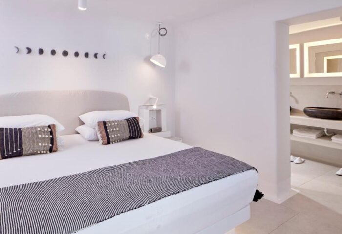 Cosmopolitan Suites gay-friendly hotel room. Minimalist design with white walls, bed with decorative pillows, and bathroom view. Elegant travel.