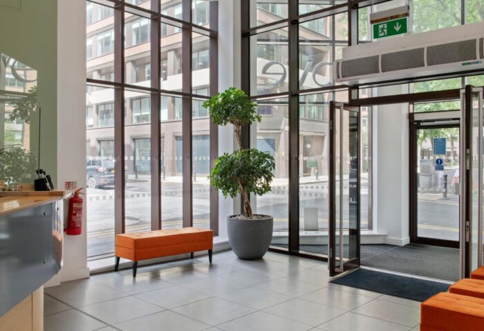 Cove Minshull Street's lobby, a gay-friendly hotel. Modern design, large windows, potted tree, and orange bench. Welcoming space for gay travelers.