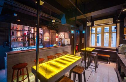 Interior view of Cube Boutique Capsule Hotel @ Chinatown's bar. Gay-friendly hotel with stools, shelves of liquor, and illuminated tables.