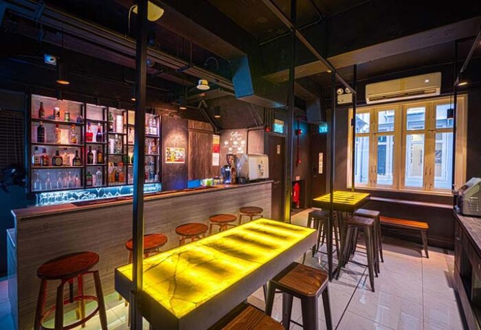 Interior view of Cube Boutique Capsule Hotel @ Chinatown's bar. Gay-friendly hotel with stools, shelves of liquor, and illuminated tables.