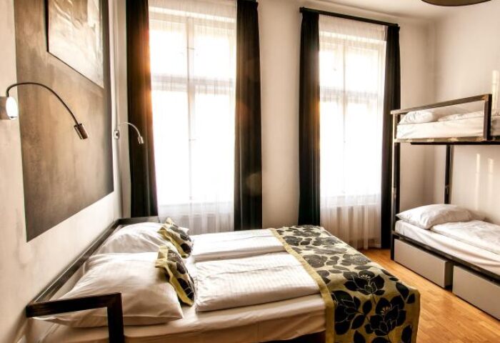 Czech Inn, gay-friendly hotel room. Double bed with floral runner, bunk beds, black curtains, and modern lamps. Ideal for gay travel.