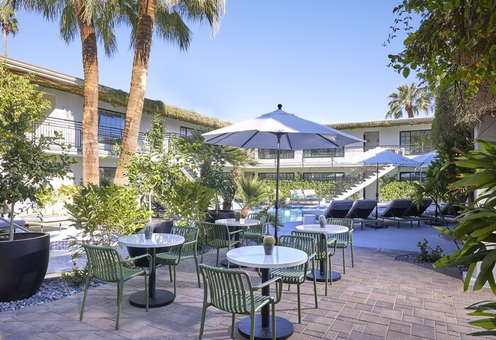 Descanso Resort, a gay-friendly men's resort. Outdoor patio with tables, chairs, palm trees, and pool. A relaxing vacation destination.