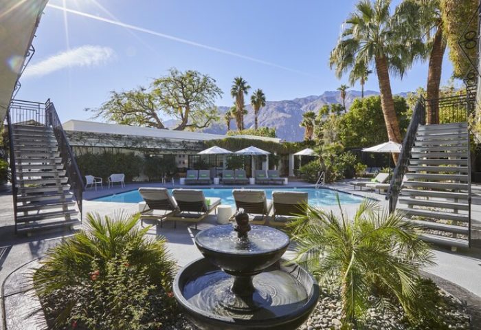 Descanso Resort, a men's gay-friendly resort. Pool view with lounge chairs, umbrellas, fountain, stairs, palm trees, and mountain backdrop.
