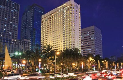 Diamond Hotel, a gay-friendly hotel, exterior view at night. The hotel is surrounded by other buildings and palm trees. Street traffic.