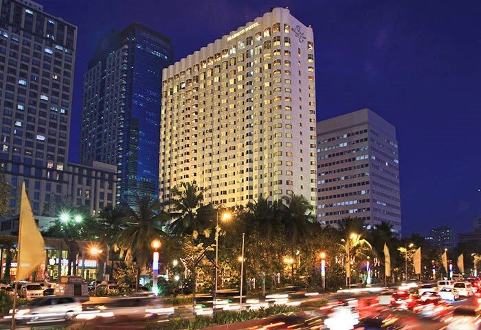 Diamond Hotel, a gay-friendly hotel, exterior view at night. The hotel is surrounded by other buildings and palm trees. Street traffic.