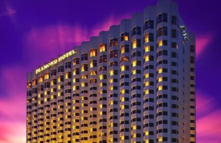 Exterior of Diamond Hotel, a gay-friendly hotel. High-rise building with many windows against a colorful sunset sky. DSC logo on top.
