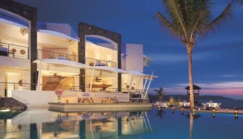 Dreams Vallarta Bay Resort & Spa all-inclusive, gay-friendly hotel. Poolside bar and dining at dusk. Palm tree, ocean view. Luxury travel.