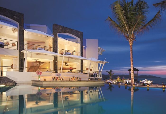 Dreams Vallarta Bay Resort & Spa all-inclusive, gay-friendly hotel. Poolside bar and dining at dusk. Palm tree, ocean view. Luxury travel.