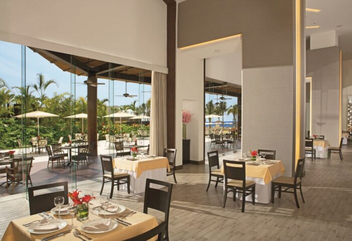 Dreams Vallarta Bay Resort & Spa all-inclusive dining. Restaurant interior with tables set, view to outdoor patio. Gay-friendly hotel.