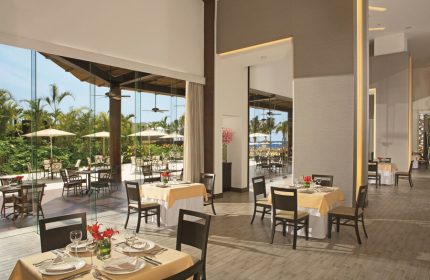 Dreams Vallarta Bay Resort & Spa all-inclusive dining. Restaurant interior with tables set, view to outdoor patio. Gay-friendly hotel.