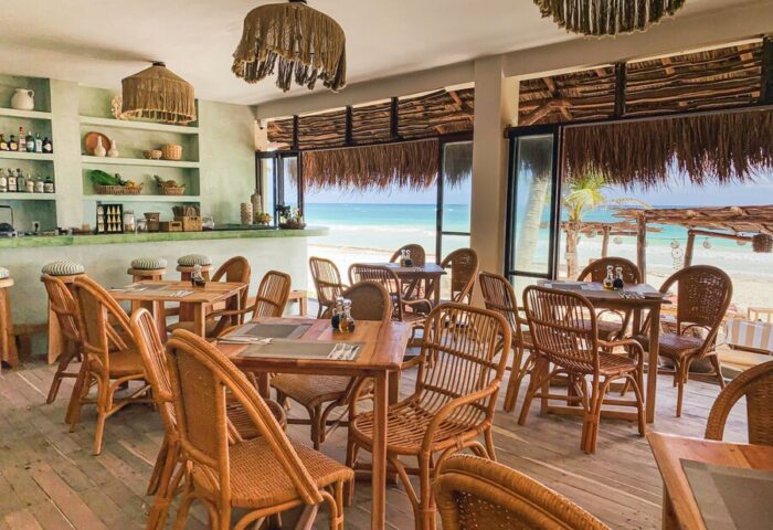 Dune Boutique Hotel's dining area in the party zone. Gay-friendly hotel with beach views, wooden tables, and woven chairs. Tropical decor.