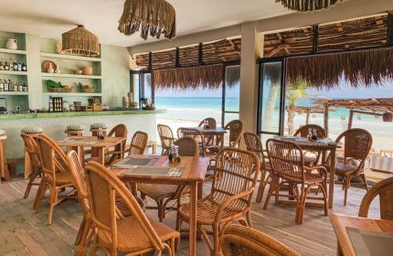 Dune Boutique Hotel's dining area in the party zone. Gay-friendly hotel with beach views, wooden tables, and woven chairs. Tropical decor.