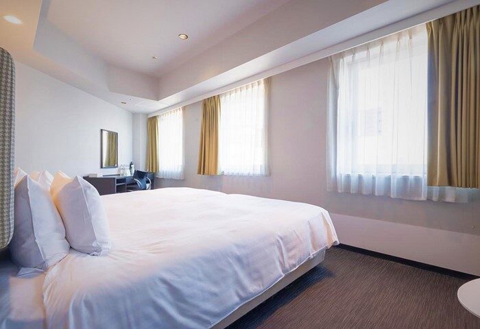 E-Hotel Higashi Shinjuku Tokyo room. Gay-friendly hotel featuring a bed with white linens, desk, chair, and windows with sheer and gold curtains.