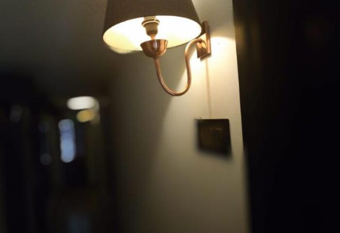 Illuminated wall lamp at East Hotel, a gay-friendly hotel. The lamp has a dark shade and a copper-colored arm, providing warm lighting.