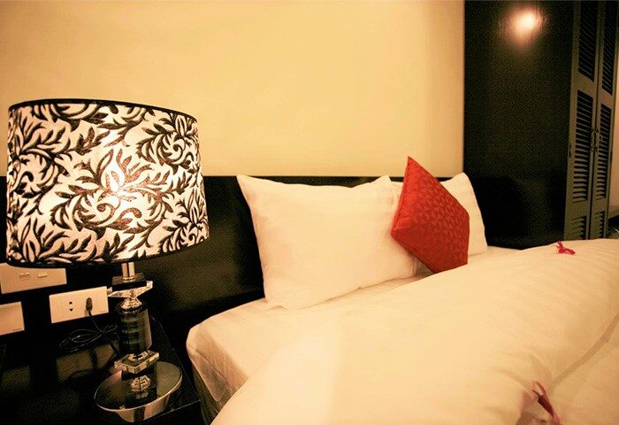 Eclipse Legend Hotel bedroom. Gay-friendly hotel room with a lamp on a bedside table, white sheets, and a red decorative pillow.