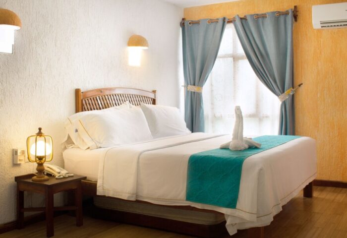 Eco-hotel El Rey del Caribe room. Gay-friendly hotel with bed, swan towel art, blue curtains, and bedside table. Gay travel lodging.