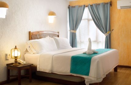 Eco-hotel El Rey del Caribe room. Gay-friendly hotel with bed, swan towel art, blue curtains, and bedside table. Gay travel lodging.