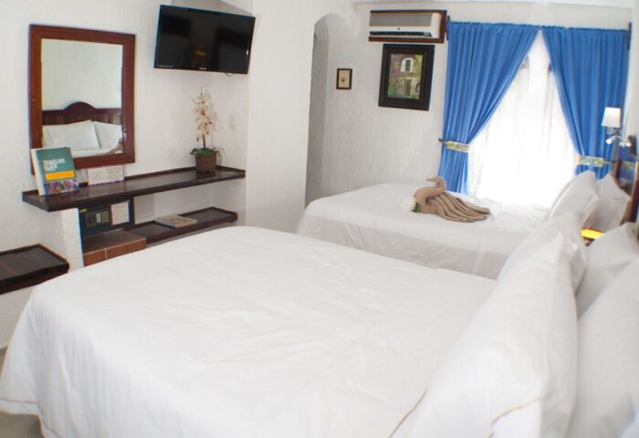 Eco-hotel El Rey del Caribe gay-friendly room. Two beds with white linens, blue curtains, TV, safe. Gay travel in a comfortable hotel.
