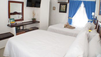 Eco-hotel El Rey del Caribe gay-friendly room. Two beds with white linens, blue curtains, TV, safe. Gay travel in a comfortable hotel.
