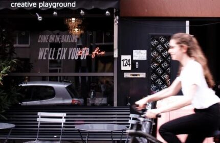Ecomama Hotel's exterior, a gay-friendly hotel. A woman rides a bike past the Creative Playground cafe. We'll fix you up neon sign.