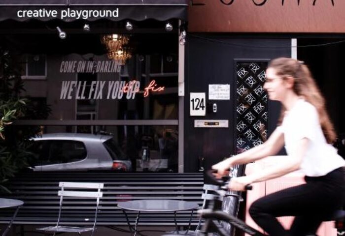 Ecomama Hotel's exterior, a gay-friendly hotel. A woman rides a bike past the Creative Playground cafe. We'll fix you up neon sign.