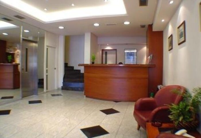 Lobby of a gay-friendly economy hotel. Reception desk, seating area, and elevator. Tile floor with black accents. Clean and well-lit.