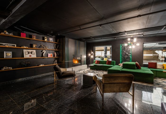 Empire Suites gay-friendly hotel lounge. Dark interior with a fireplace, green sofas, and shelves with decorative objects. Modern design.