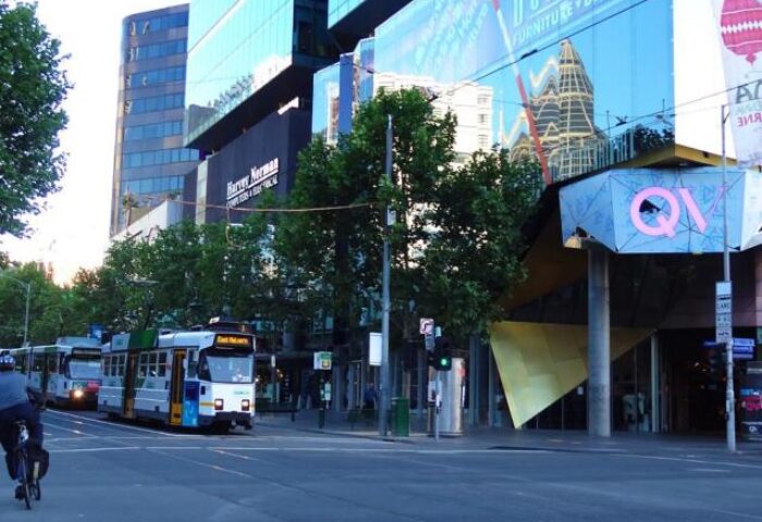 Street view of Essence Hotel Carlton, a gay-friendly hotel. Trams on the street, modern buildings, and a cyclist. Carlton, Australia.