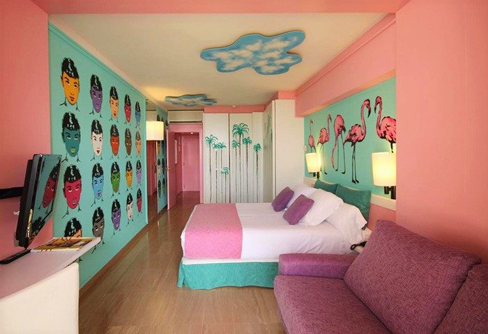 Estela Barcelona gay-friendly hotel room. Colorful walls with flamingo and face art. Double bed, sofa, and cloud-shaped ceiling decor.
